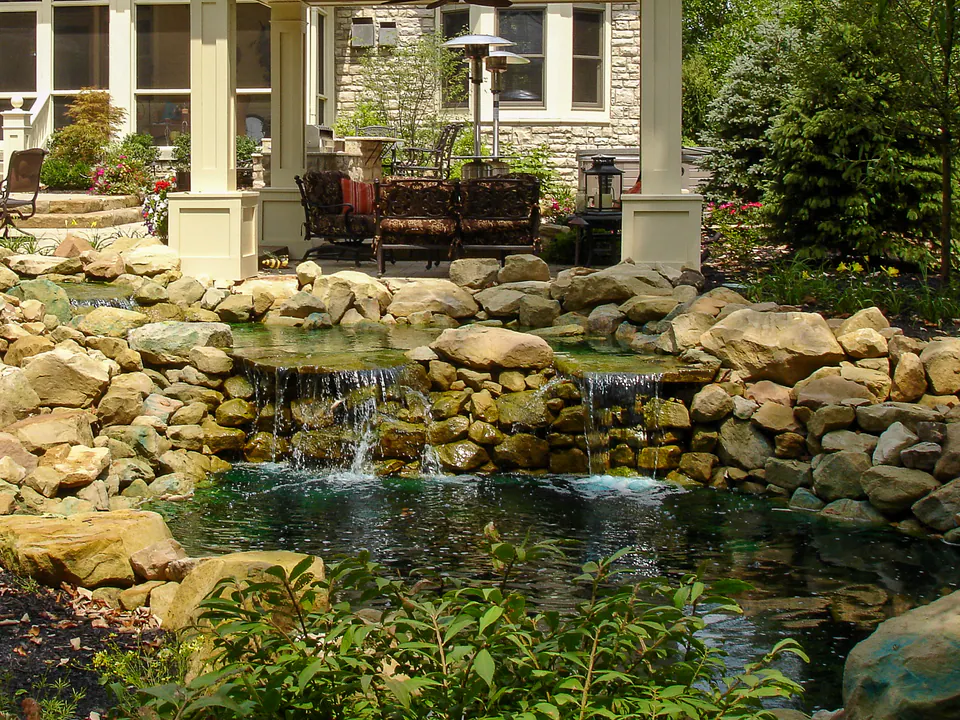 Backyard pond with natural boulder edge, small waterfall, patio seating, and house beyond